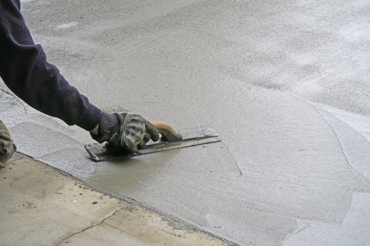 A gloved hand smoothing a fresh concrete surface with a steel trowel.