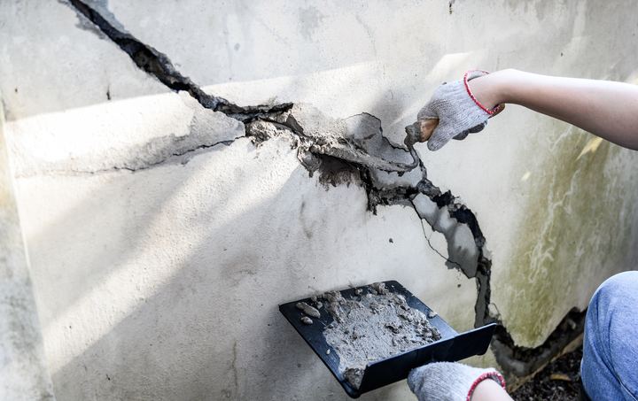A gloved hand troweling mortar into a large crack in a concrete foundation wall.