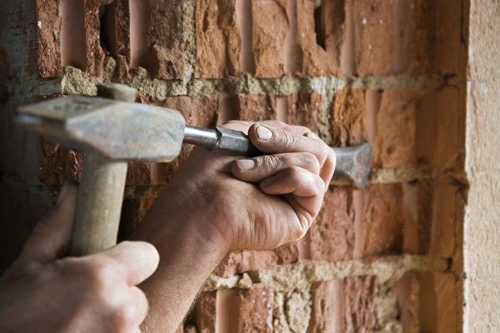 Hands using a hammer and chisel to chip out old mortar from a brick wall.
